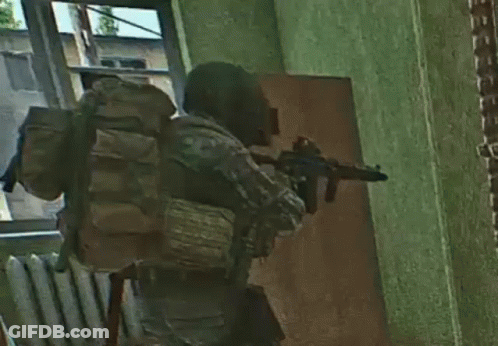 A Soldier With A Backpack Is Holding A Gun In A Room With A Door . GIF