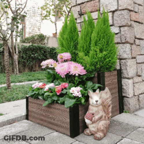 A Squirrel Statue Sits Next To A Wooden Planter Filled With Flowers And Plants GIF
