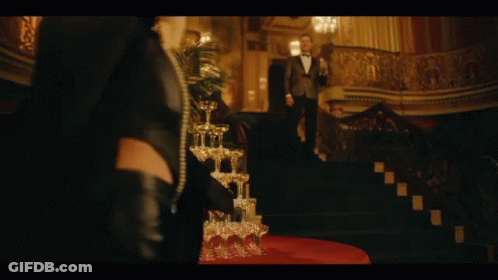 A Stack Of Champagne Glasses On A Table With A Woman Standing In The Background GIF