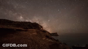 A Starry Night Sky Over A Body Of Water With A Cliff In The Foreground GIF