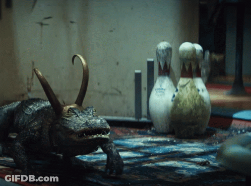 A Statue Of A Crocodile With A Crown On Its Head Stands Next To Bowling Pins GIF