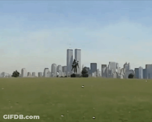 A Statue Of A Man Carrying Another Man In Front Of The Twin Towers Of The World Trade Center GIF