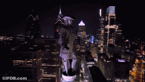 A Statue Of A Man In A Hat Stands On Top Of A City Skyline At Night GIF