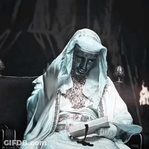 A Statue Of A Man In A White Robe Sits In A Chair Reading A Book GIF