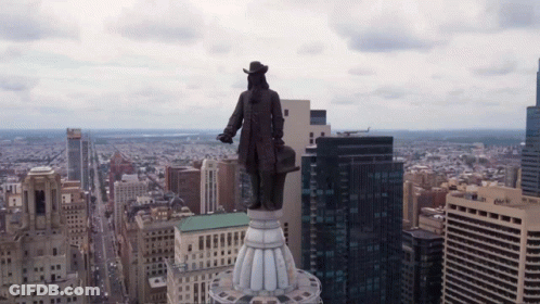 A Statue Of A Man Stands On Top Of A Building In A City With A Chase Bank Building In The Background GIF