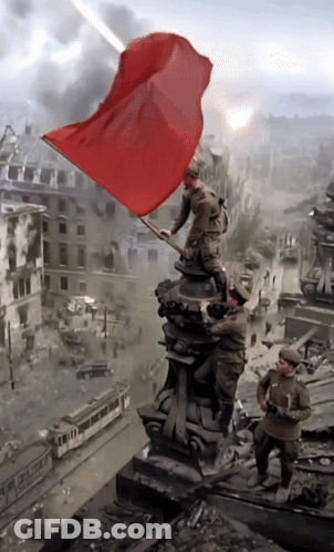 A Statue Of Soldiers Holding A Red Flag In Front Of A Destroyed City GIF