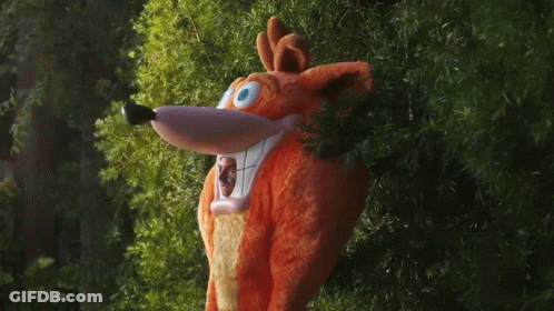 A Stuffed Animal In A Crash Bandicoot Costume Peeking Out From Behind Some Trees GIF