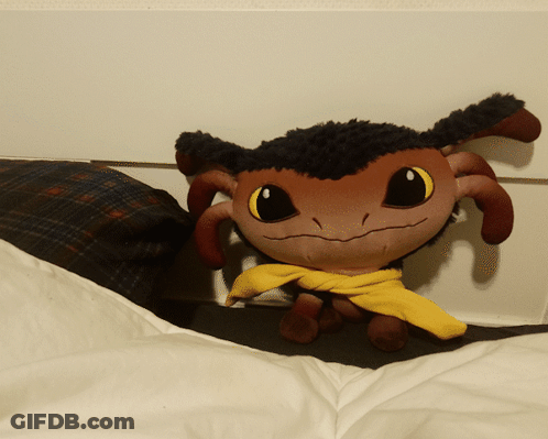 A Stuffed Animal With A Yellow Scarf Around Its Neck Is Sitting On A Bed GIF