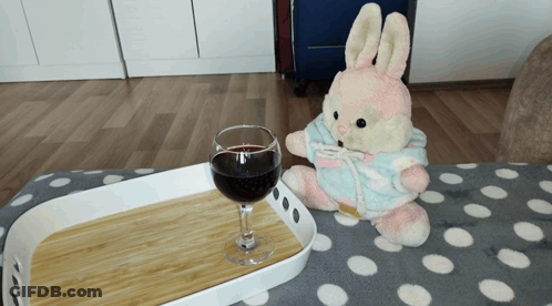 A Stuffed Bunny Is Sitting Next To A Glass Of Wine On A Tray GIF