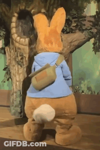 A Stuffed Bunny Is Standing Next To A Tree Wearing A Blue Jacket And A Green Bag . GIF