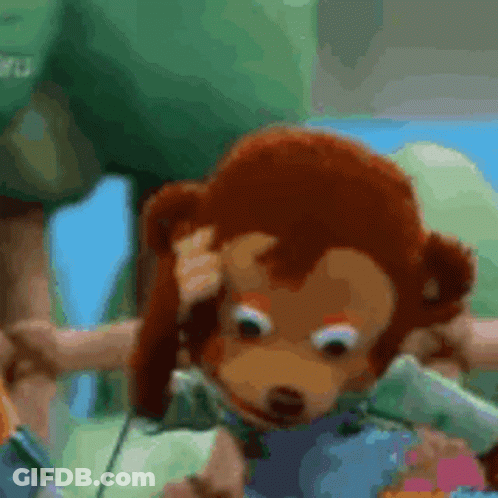 A Stuffed Monkey Is Scratching His Head While Sitting On A Green Blanket . GIF