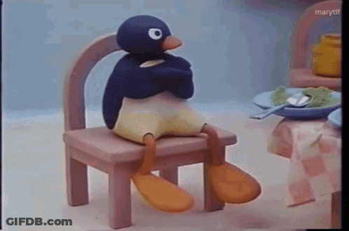 A Stuffed Penguin Is Sitting On A Wooden Chair In Front Of A Table . GIF