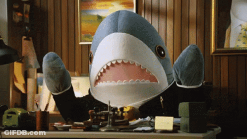 A Stuffed Shark In A Suit Is Sitting At A Desk With Its Mouth Open GIF