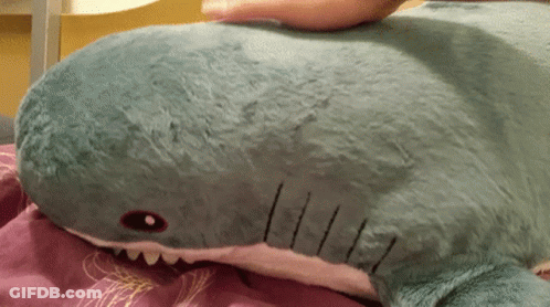 A Stuffed Shark Is Laying On A Bed With A Person 'S Hand On It GIF