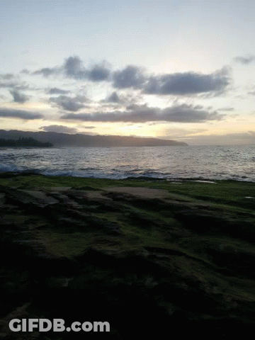 A Sunset Over A Body Of Water With A Grassy Hill In The Foreground GIF