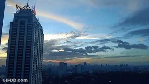 A Sunset Over A City With A Skyscraper In The Foreground That Says Shark GIF