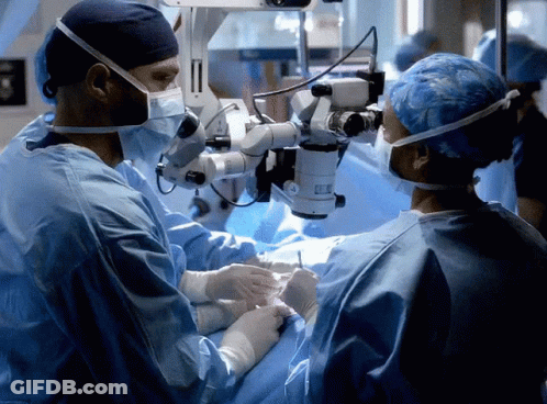 A Surgeon Is Looking Through A Microscope While Another Surgeon Looks On GIF