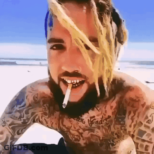 A Tattooed Man With Dreadlocks Is Smoking A Cigarette On The Beach GIF
