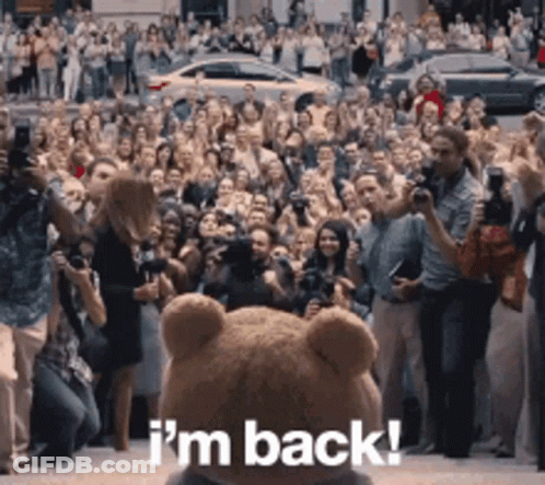 A Teddy Bear Is Standing In Front Of A Crowd That Says I 'M Back GIF