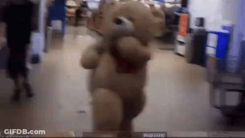 A Teddy Bear Is Walking Down A Hallway In A Store With A Person Behind It . GIF