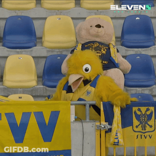 A Teddy Bear Sits Next To A Mascot In A Stadium GIF
