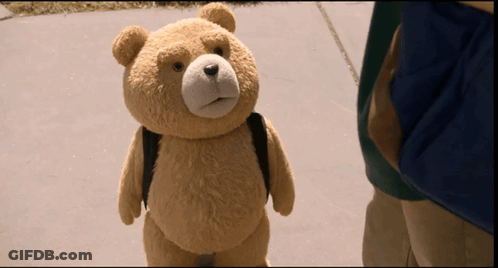 A Teddy Bear Wearing A Backpack Stands On A Sidewalk GIF