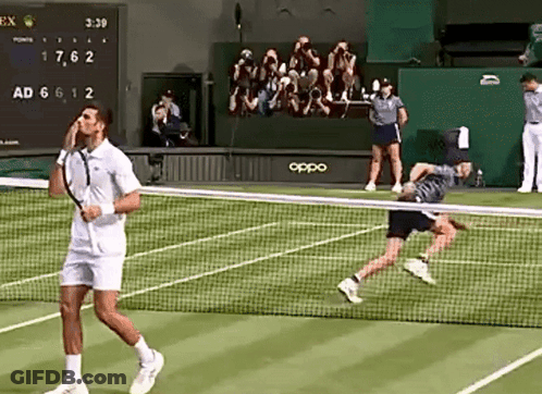 A Tennis Player Is Talking On A Cell Phone While Another Player Is Running Towards The Net . GIF