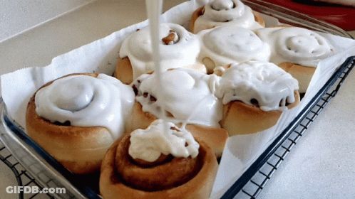 A Tray Of Cinnamon Rolls With White Frosting Being Poured On Them GIF