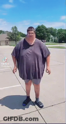 A Very Fat Man Is Standing On A Sidewalk Wearing A Gray Shirt And Sandals . GIF
