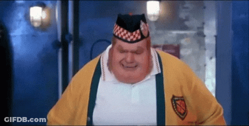 A Very Fat Man Wearing A Yellow Sweater And A Hat Is Standing In A Dark Room . GIF