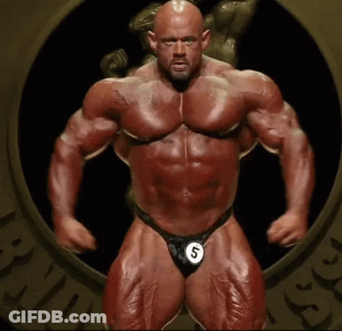 A Very Muscular Man Is Standing In Front Of A Statue Wearing A Number 5 . GIF