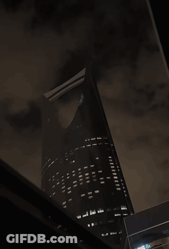 A Very Tall Building Is Lit Up At Night With A Dark Sky GIF