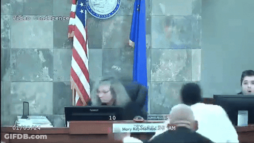 A Video Conference Is Being Held In A Courtroom With A Flag Behind The Judge GIF