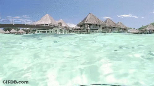 A View Of A Tropical Island From The Water With Thatched Houses In The Background GIF