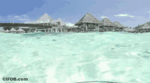 A View Of A Tropical Island From The Water With Thatched Huts In The Background GIF
