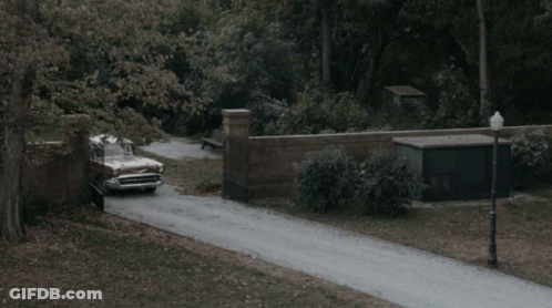 A Vintage Car Is Driving Down A Road With A Brick Wall In The Background GIF