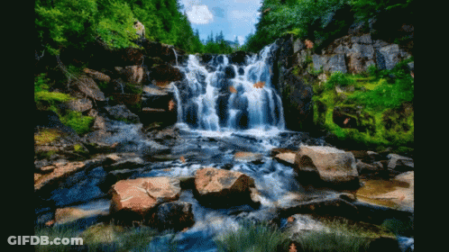 A Waterfall Surrounded By Rocks And Trees With A Person Standing In The Background GIF