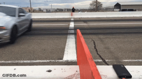 A White Car Is Driving Down A Road With A Red Barrier In The Foreground GIF