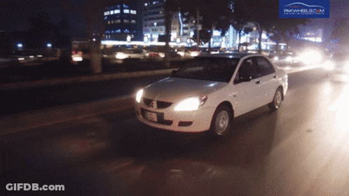 A White Car Is Driving Down A Street At Night With A Pakwheels.com Logo Behind It GIF