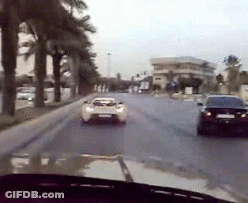 A White Car Is Driving Down A Street Next To A Black Car . GIF
