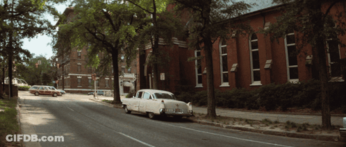 A White Car Is Parked On The Side Of The Road In Front Of A Red Brick Building GIF