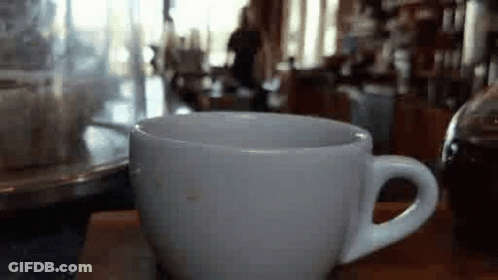 A White Cup Of Coffee Is Sitting On A Wooden Table In A Cafe . GIF