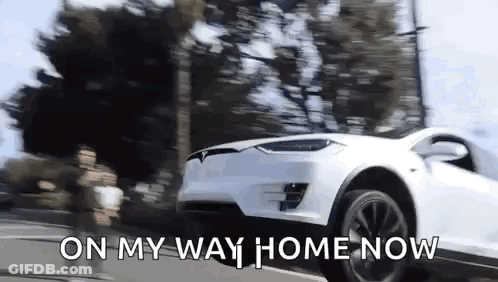 A White Tesla Model X Is Driving Down A Street With The Words `` On My Way Home Now '' Written On The Bottom . GIF