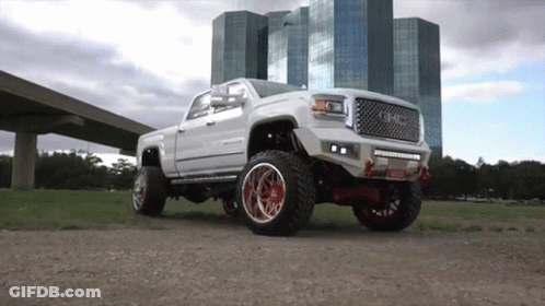 A White Truck With Red Wheels Is Parked In A Field In Front Of A Building . GIF