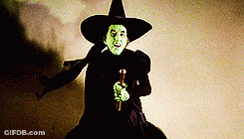 A Witch With Green Eyes And A Black Hat Is Holding A Cane . GIF