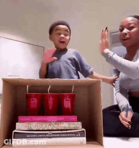 A Woman And A Boy Are Playing With A Cardboard Box Containing A Stack Of Books Including One By Oscar De La Rente GIF
