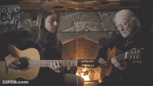 A Woman And An Older Man Are Playing Guitars In Front Of A Fireplace GIF