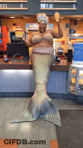 A Woman Dressed As A Mermaid Holds A Drink In Front Of A Pop Corn Machine GIF