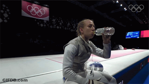 A Woman Drinking Water From A Bottle In Front Of A Sign That Says Olympic Rings GIF