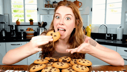 A Woman Eating A Chocolate Chip Cookie From A Plate Of Cookies GIF